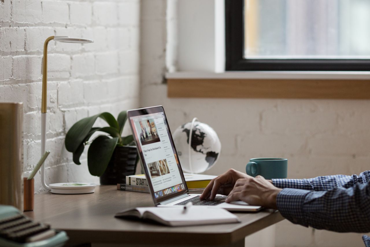 A tidy, modern workspace showing a person working on a laptop at a desk decorated with a globe, lamp, and plant.